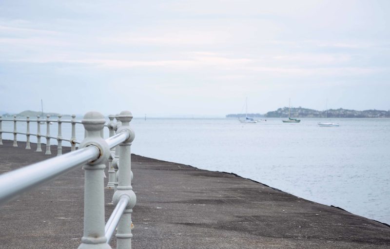White metal railing along a waterfront promenade overlooking a calm bay with distant sailboats and a hazy coastline